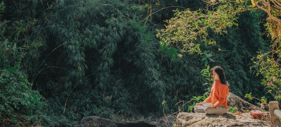 Woman meditating by a serene riverside surrounded by lush greenery on a sunny day.